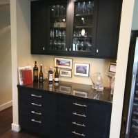 This wine bar features cabinets that match the kitchen cabinets, but in a black finish, and a tall wine storage unit to the right.